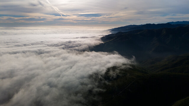 Aerial view of a blanket of clouds meeting the rugged, dark mountains under a sky streaked with contrails, Malibu, California, United States.