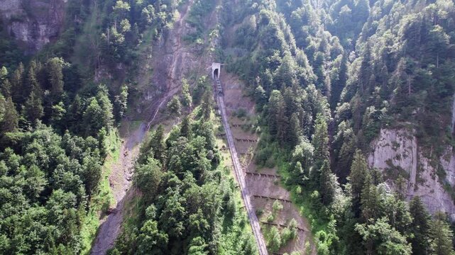 4K aerial drone view of the Stoosbahn, the world's steepest funicular, ascending a lush forested mountainside. Showcasing Swiss engineering and scenic alpine transport to the car-free Stoos village.