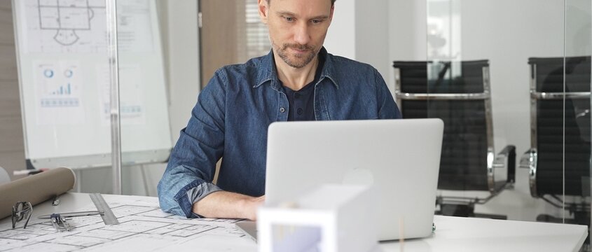 Male architect wearing denim shirt working on laptop in modern office, analyzing blueprints and architectural plans on desk, designing new building construction project
