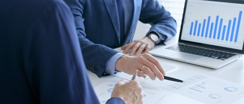 Business colleagues or partners are discussing financial data on a laptop and printed documents while analyzing charts and market trends at office table. Businesspeople and finance concept