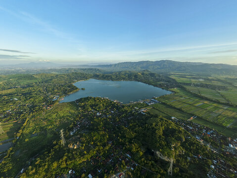 Aerial view of the vast, tranquil Rawa Jombor lake reflecting the clear blue sky, embraced by lush green hills and distant mountains, Klaten, Jawa Tengah, Indonesia.