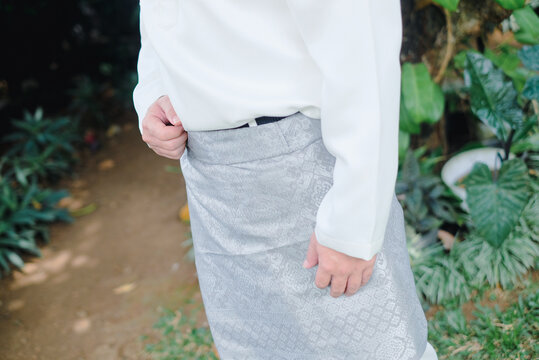 Person wearing a traditional grey patterned sarong (sampin) and white baju melayu shirt stands outdoors for a cultural event or celebration like Eid al-Fitr.
