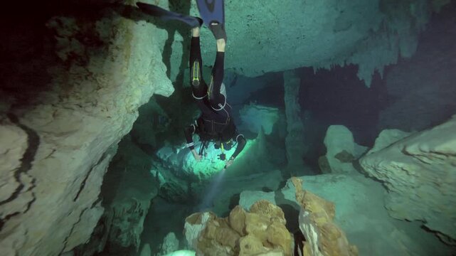 Diver swims between limestone columns and cave walls with visible sediment floor in Cenote Dos Ojos, Quintana Roo, Mexico