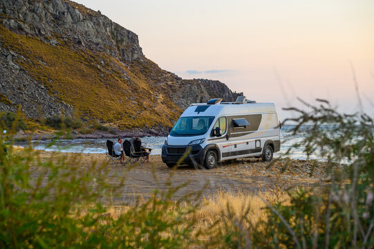 Family relax in folding chairs next to their modern campervan on a rugged beach in Limnos, Greece, enjoying a sunset view of the Aegean Sea during an off-grid motorhome road trip.