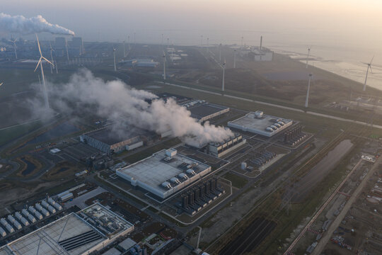 Aerial view of large scale data center campus processing artificial intelligence, with cooling towers emitting steam, powered by surrounding wind turbines in the netherlands