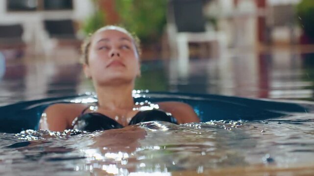 Asian woman floating in pool closeup portrait with bun hairstyle, urban resort backdrop, lights glittering on water, calm confident expression, wellness and leisure vibe, swim practice