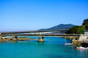 Fototapeta premium Coastal bridge with small fishing boats on clear blue water in Shikoku Japan with mountain backdrop and calm seaside atmosphere