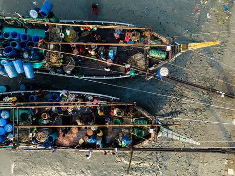 Aerial view of fishing boats laden with blue barrels and busy with people, docked on the muddy shores, Chattogram, Chittagong Division, Bangladesh.