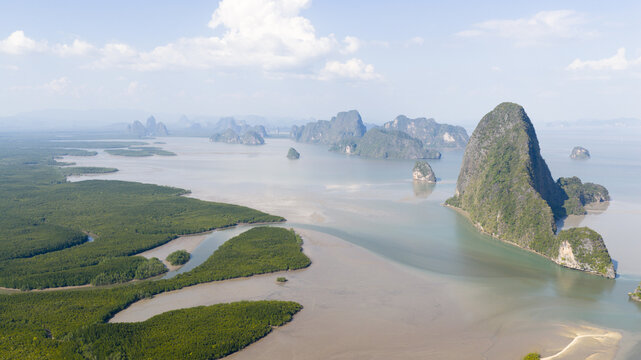 Aerial view of limestone karsts rising from the calm sea and lush mangrove forests under a bright sky in Phang Nga, Thailand.