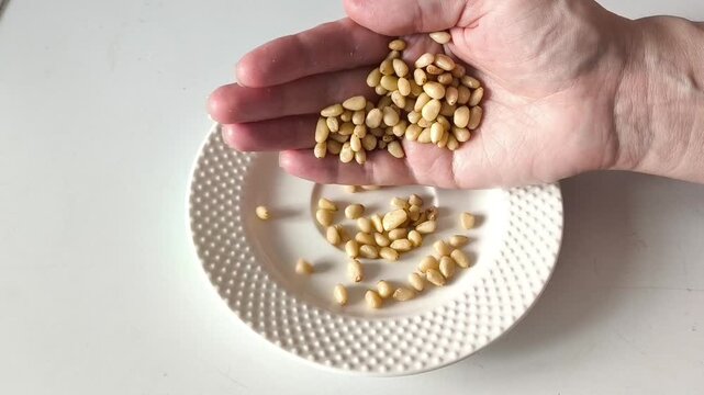 A handful of pine nuts in his hand. A woman's hand pours a kernel of cedar nuts into a white plate on the table