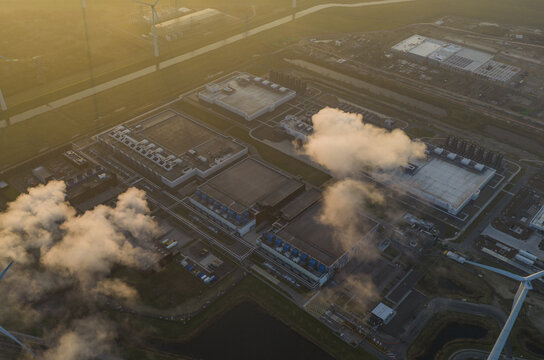 Aerial view of large data center facilities emitting steam, surrounded by wind turbines, symbolizing modern technology, cloud computing, and renewable energy