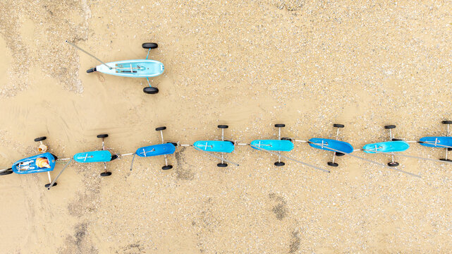 Aerial view of a line of beach yachts resting on the beach, their vibrant blue bodies contrasting with the tan sand, Fort-Mahon-Plage, Hauts-de-France, France.