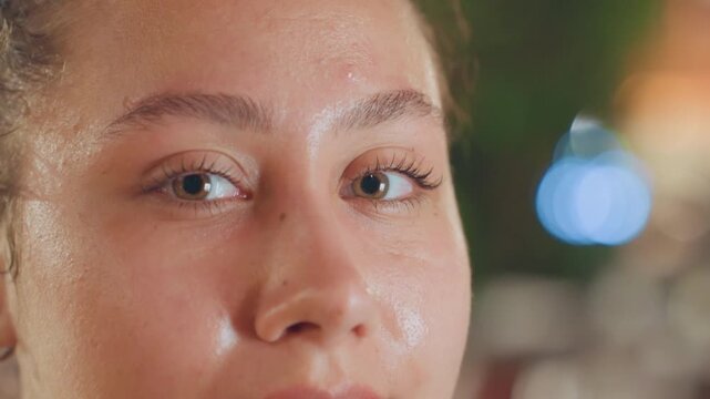 Closeup woman reflective gaze island lights, dew on skin with shimmering highlights, soft film grain and bokeh background suggest intimate seaside dinner or poolside evening, calm contemplative