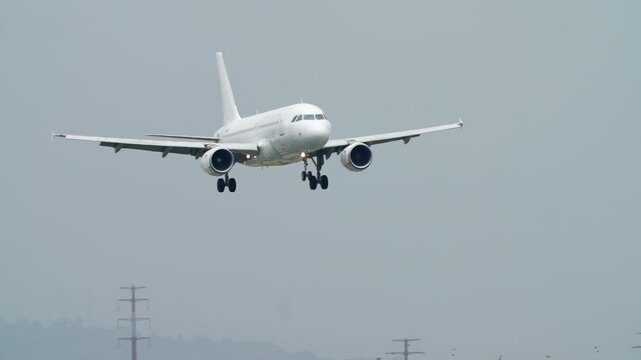 A white, unmarked passenger jet descends for landing. The aircraft flies closer to the camera with landing gear down over a city on a cloudy, hazy day. A view of final approach before touchdown