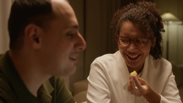 Tracking shot of young African American woman talking to her husband and feeding him with grape while he working on laptop