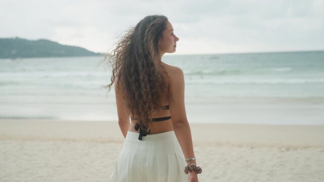 Lady watches far marine horizon. Female with flowing hair surveys expansive marine landscape at distance. Woman contemplating endless ocean scenery from her seated position by quiet shoreline