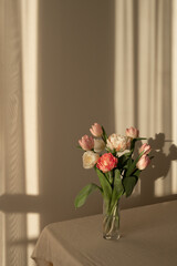 Wase with tulip flowers bouquet on table with neutral beige linen tablecloth and empty wall background with abstract natural warm evening sunlight shadows, floral home interior decor