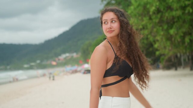 Curly woman smiling island beach shore, playful glance and hair toss, black bikini top with white shorts, distant green hills and cloudy sky, bracelets and relaxed posture, joyful energy, candid