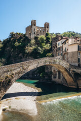 Obraz premium Historic stone bridge Ponte Vecchio over the clear waters of the Nervia river with the ruins of Doria Castle on the hill and traditional houses, sunny day in Dolceacqua, Liguria, Italy