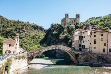 Naklejka premium Historic stone bridge Ponte Vecchio over the clear waters of the Nervia river with the ruins of Doria Castle on the hill and traditional houses, sunny day in Dolceacqua, Liguria, Italy