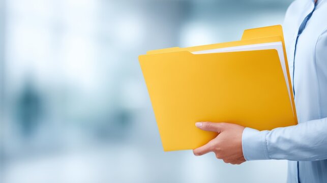 Business professional in a light blue shirt holds a stack of yellow folders in a modern office environment with blurred background elements suggesting a corporate setting