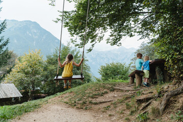 Fototapeta premium Little girl sitting together on a wooden tree swing overlooking a mountain valley.
