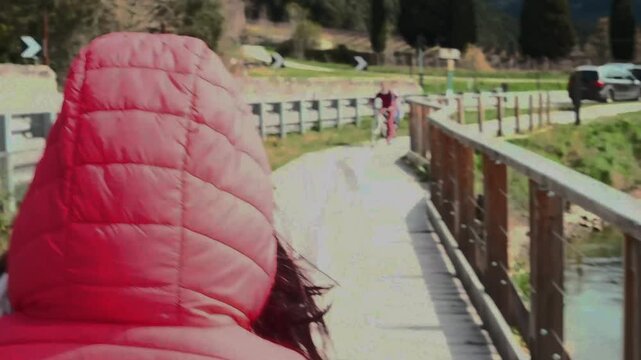 A woman, photographed from behind, walks on a walkway overlooking Lake Tublin in Trentino, Northern Italy. The woman has long black hair and wears a fuchsia down jacket