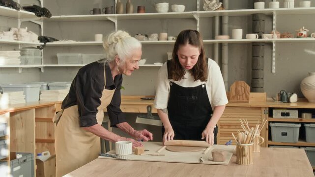 Young woman learning to work with clay while elderly craftswoman teaching her at workbench in modern pottery studio