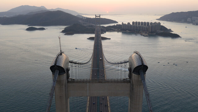 Aerial view of the Tsing Ma Bridge at sunset with traffic crossing the water between the towers towards distant islands and high-rise buildings in Hong Kong, China.