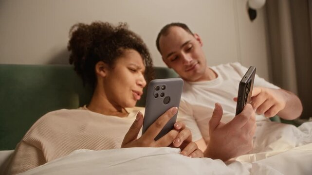 Handheld shot of young African American woman and young man using smartphones while resting in bed
