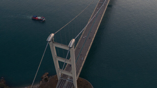 Aerial view of the Tsing Ma Bridge with a cargo ship sailing on the blue water and traffic crossing the suspension bridge in Hong Kong, China.
