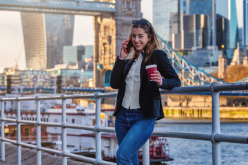 Fototapeta premium A beautiful, young woman talks on her moblile phone and holding a coffee cup in front of the city skyline of London as a business concept