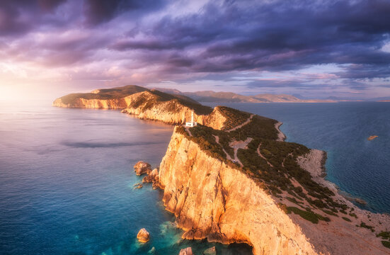 Lighthouse on the mountain peak at colorful sunset in summer. Aerial view. Beautiful lighthouse on the rock, sea and moody sky with purple clouds at dusk. Top view of Cape Lefkada, Greece. Landscape