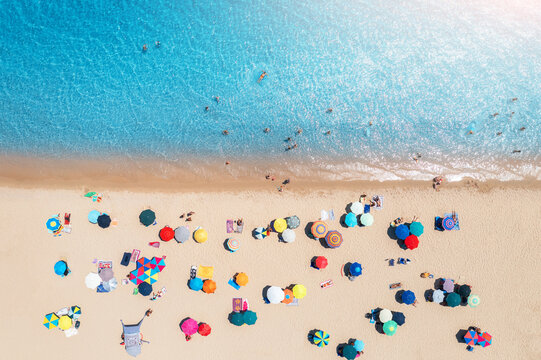 Aerial view of colorful umbrellas on sandy beach, swimming people in blue sea at sunset in summer in Sardinia, Italy. Tropical landscape with turquoise transparent water. Travel and vacation. Top view
