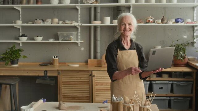 Portrait shot of elderly craftswoman smiling for camera while working on laptop in her moden pottery studio