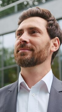 Close-up of a bearded businessman face, smiling confidently while looking to the side outdoors in an urban business environment. People and business concept. Slow motion
