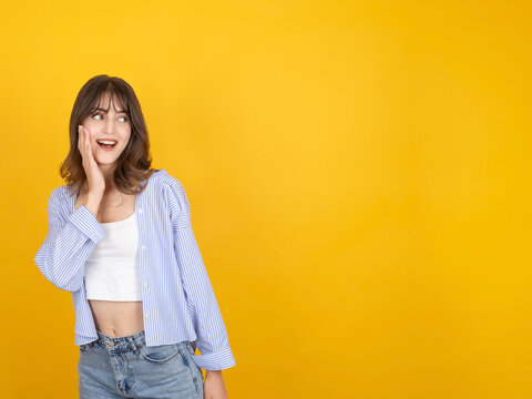 Excited caucasian woman touching her cheek with surprised smile, looking aside with curiosity, wearing striped shirt and denim, isolated on bright yellow studio background with copy space.