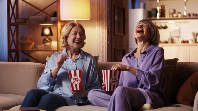 Two happy senior women friends watching comedy show on TV, eating popcorn