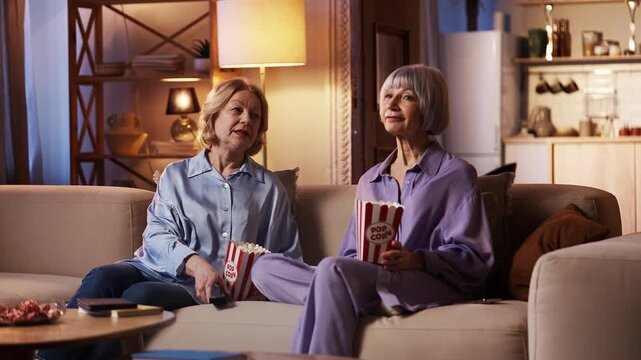 Smiling sisters in their 60s watching movie and eating popcorn, leisure time