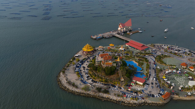 Aerial view of the Wat Ko Loi temple complex on a small peninsula featuring traditional Thai architecture, a long pier, and numerous fish farms in the sea at Si Racha, Chon Buri, Thailand.