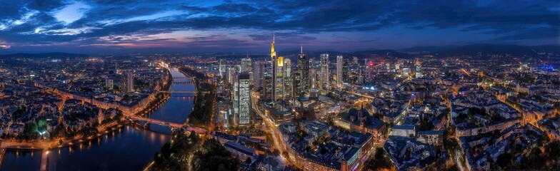 Fototapeta premium Aerial panorama of a city at dusk, with illuminated buildings, a river, and a colorful sky