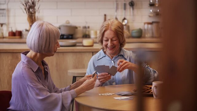 Smiling senior woman playing card game with female friend at home, fun
