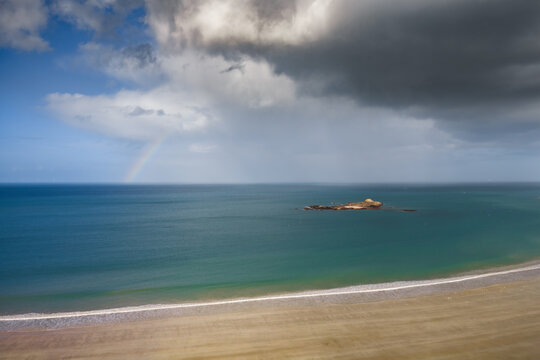 Aerial view of the sandy beach and Ilot Saint-Michel with a rainbow under dark storm clouds in Erquy, Brittany, France.