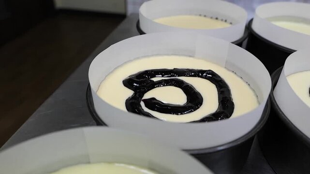 A close-up shot shows a professional pastry chef, wearing white gloves, using a black piping bag to create a decorative swirl pattern with dark blueberry jam or sauce on top of several individual chee