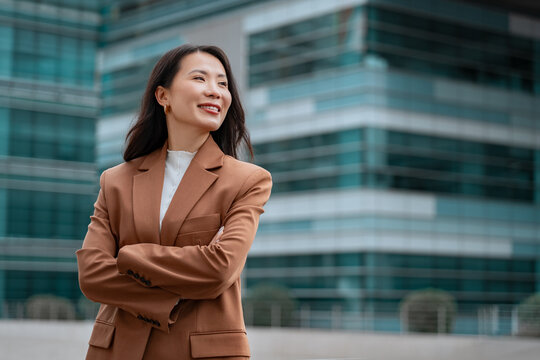 Portrait Asian mature businesswoman standing arms crossed outdoors looking dreaming aside at copy space. Middle-aged business woman entrepreneur female executive in suit smiling at office skyscraper
