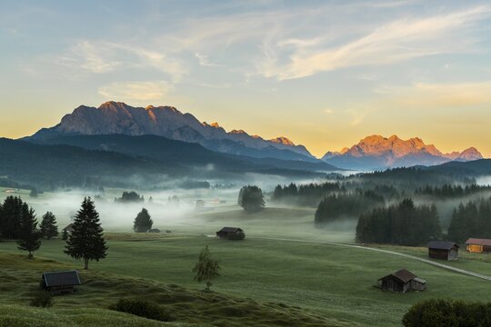 Mountain meadows with small huts in light ground fog and Zugspitz massif in the background at sunrise, Kr&uuml;n, Bavaria, Germany