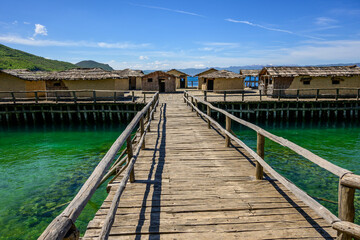 Fototapeta premium A long wooden boardwalk leads directly into the reconstructed Bay of Bones prehistoric stilt village, with transparent turquoise Lake Ohrid water glistening under a blue sky in North Macedonia.