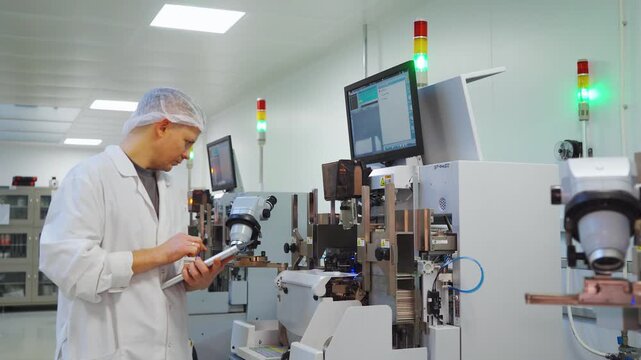 Technician inspecting microchip assembly line in sterile cleanroom using tablet to monitor semiconductor production with highprecision equipment, indicator lights and automated feeders