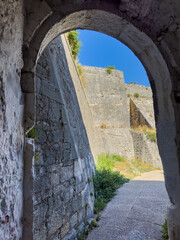 Fototapeta premium Stone arch frames the weathered ramparts of the New Fortress in Corfu, Greece. Quiet path, blue sky.