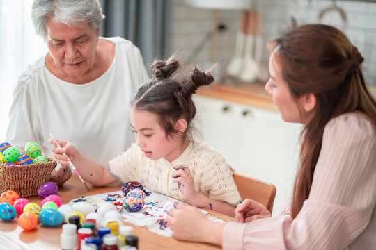 Three generations gather around a table full of colorful Easter eggs and paint, each carefully decorating their own egg in a joyful and creative family tradition.